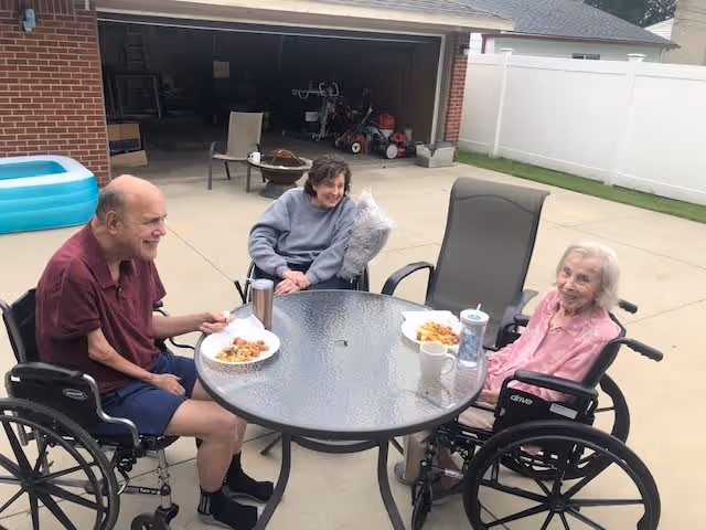 Three elderly people sitting in wheelchairs around a round glass outdoor table on a concrete patio. They are eating food from white plates and appear to be enjoying a meal together. Behind them is a garage with an open door and a white fence enclosing the area.
