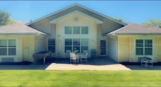 Exterior view of a single-story building with a central peaked roof section and two wings on either side. The building has multiple windows and a patio area with a table and chairs in the center. The foreground features a well-maintained green lawn under a clear blue sky.