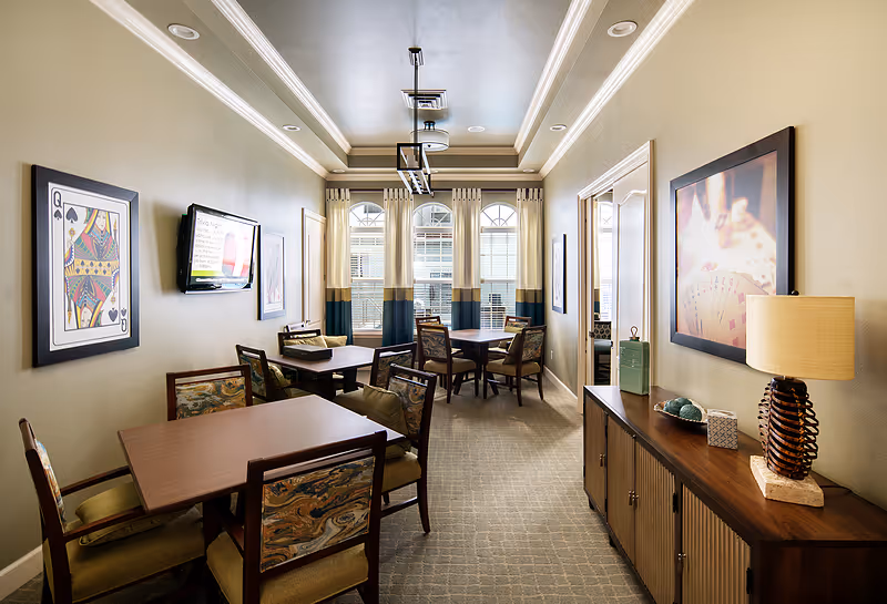 A well-lit interior room with several wooden tables and cushioned chairs arranged for small group seating. The walls are decorated with framed artwork, including a large playing card and a picture of playing cards. A flat screen TV is mounted on the left wall. A wooden sideboard with decorative items and a lamp is on the right. Large windows with curtains allow natural light into the room.