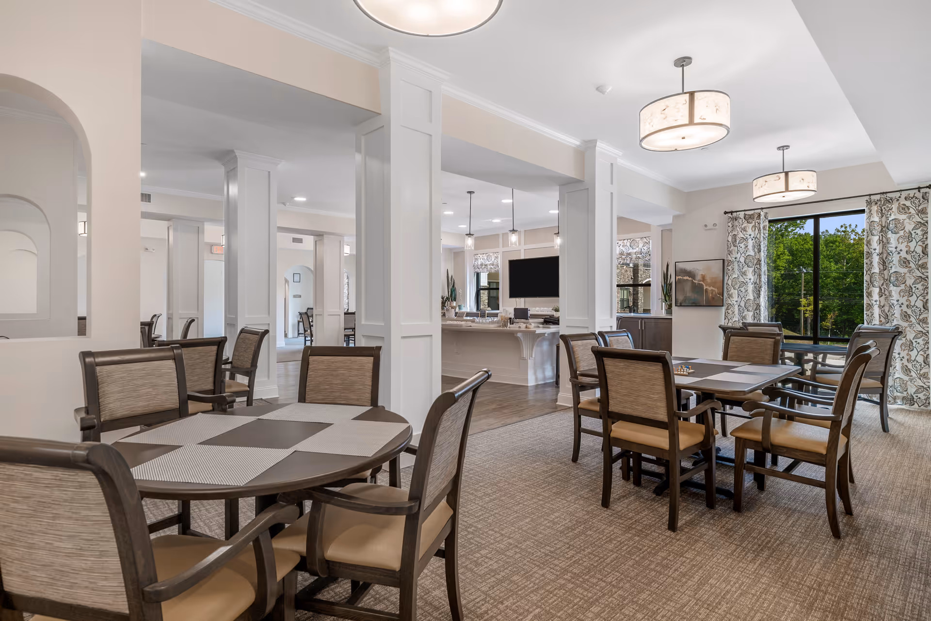 A bright and spacious dining area in a senior living facility featuring multiple tables with chairs arranged around them. The room has large windows with patterned curtains allowing natural light to enter, modern light fixtures on the ceiling, and a view into an adjacent kitchen area with a large TV mounted on the wall.