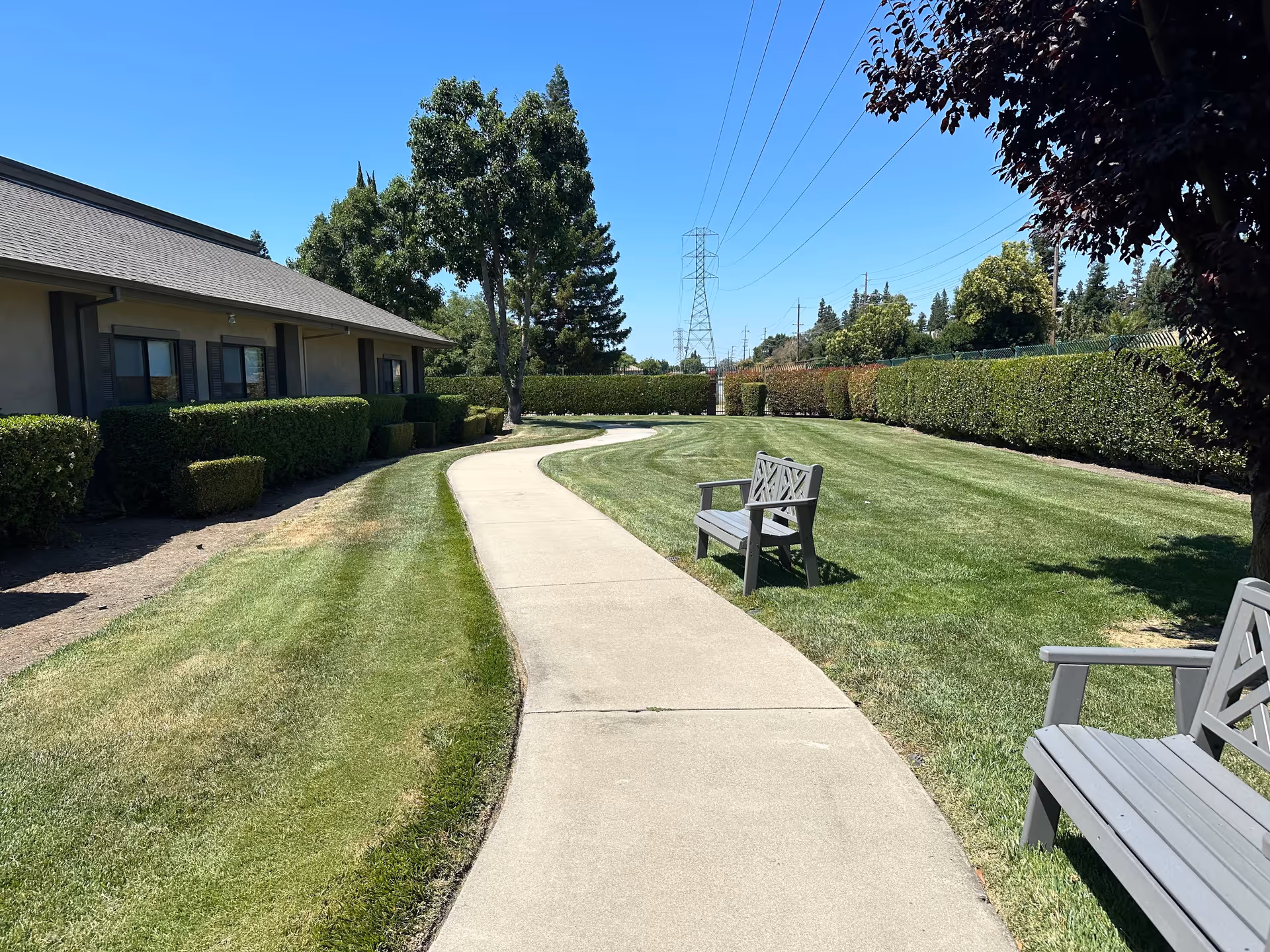 A paved walkway curves through a well-maintained grassy outdoor area with two gray benches on either side. There are trimmed hedges and trees lining the path, and a building with windows and a sloped roof is visible on the left. The sky is clear and blue.