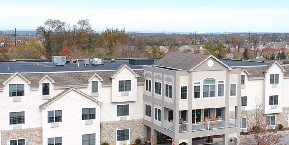 Exterior view of a multi-story senior living facility named The Willow, featuring a combination of stone and siding on the facade, multiple windows, and a covered balcony area with chairs. Trees with autumn foliage and a distant residential area are visible in the background.