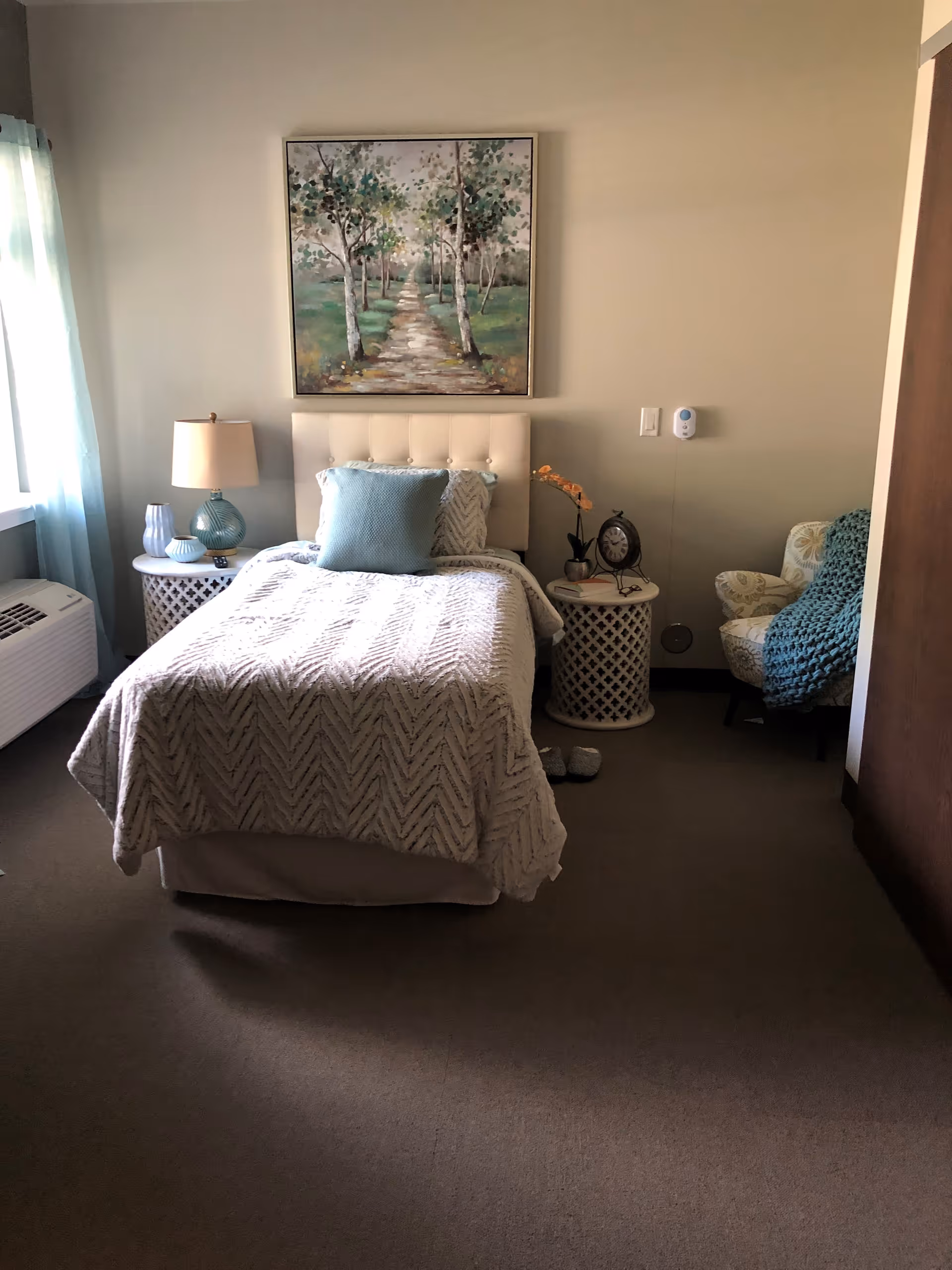 A cozy bedroom with a single bed covered in a textured white blanket and a blue pillow. There are two round white side tables on either side of the bed, one with a lamp and decorative items, the other with a clock and a small plant. A painting of a tree-lined path hangs above the bed. A cushioned armchair with a blue knitted throw is positioned in the corner. Light enters through a window with sheer blue curtains on the left side.