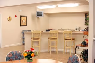 Interior view of a senior living facility common area with a bar counter and three wooden bar stools. There is a small wall-mounted TV above the counter. In the foreground, a round table with floral-patterned chairs and a small flower arrangement is visible. The walls are light-colored, and there are framed pictures and some plants around the room.
