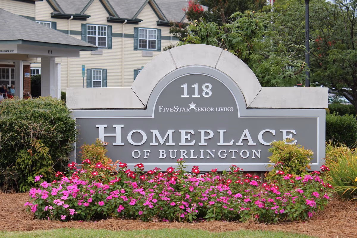 Outdoor view of a sign for Home Place of Burlington, a Five Star Senior Living facility, surrounded by colorful flowers and greenery with a building in the background.