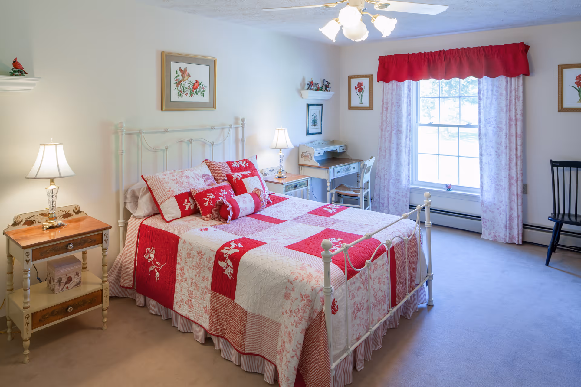 A bright and cozy bedroom with a white metal bed frame and a red and white patchwork quilt. The bed is adorned with multiple matching pillows. There is a wooden nightstand with a lamp on the left side of the bed. A small desk with a chair is positioned near a large window with red valance and floral curtains. The walls are decorated with framed floral artwork and small shelves with decorative items.