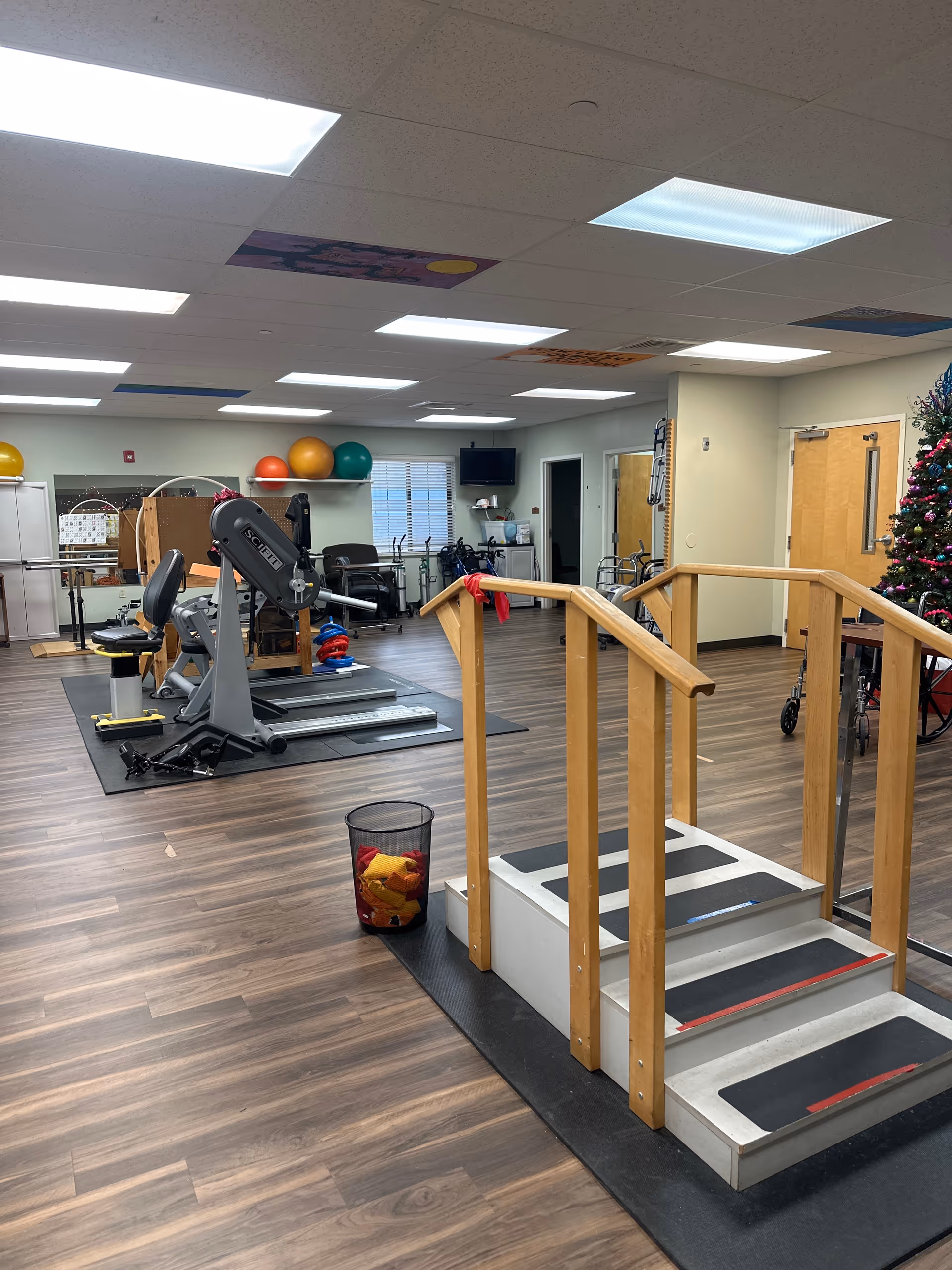 A spacious rehabilitation room with wooden flooring and bright overhead lights. The room contains exercise equipment including a seated exercise machine, therapy balls on a shelf, parallel bars, and a small set of stairs with handrails for physical therapy. There is a Christmas tree decorated with ornaments in the corner near a door, and a trash bin with colorful cloths inside. The room has a window with blinds and a wall-mounted TV.