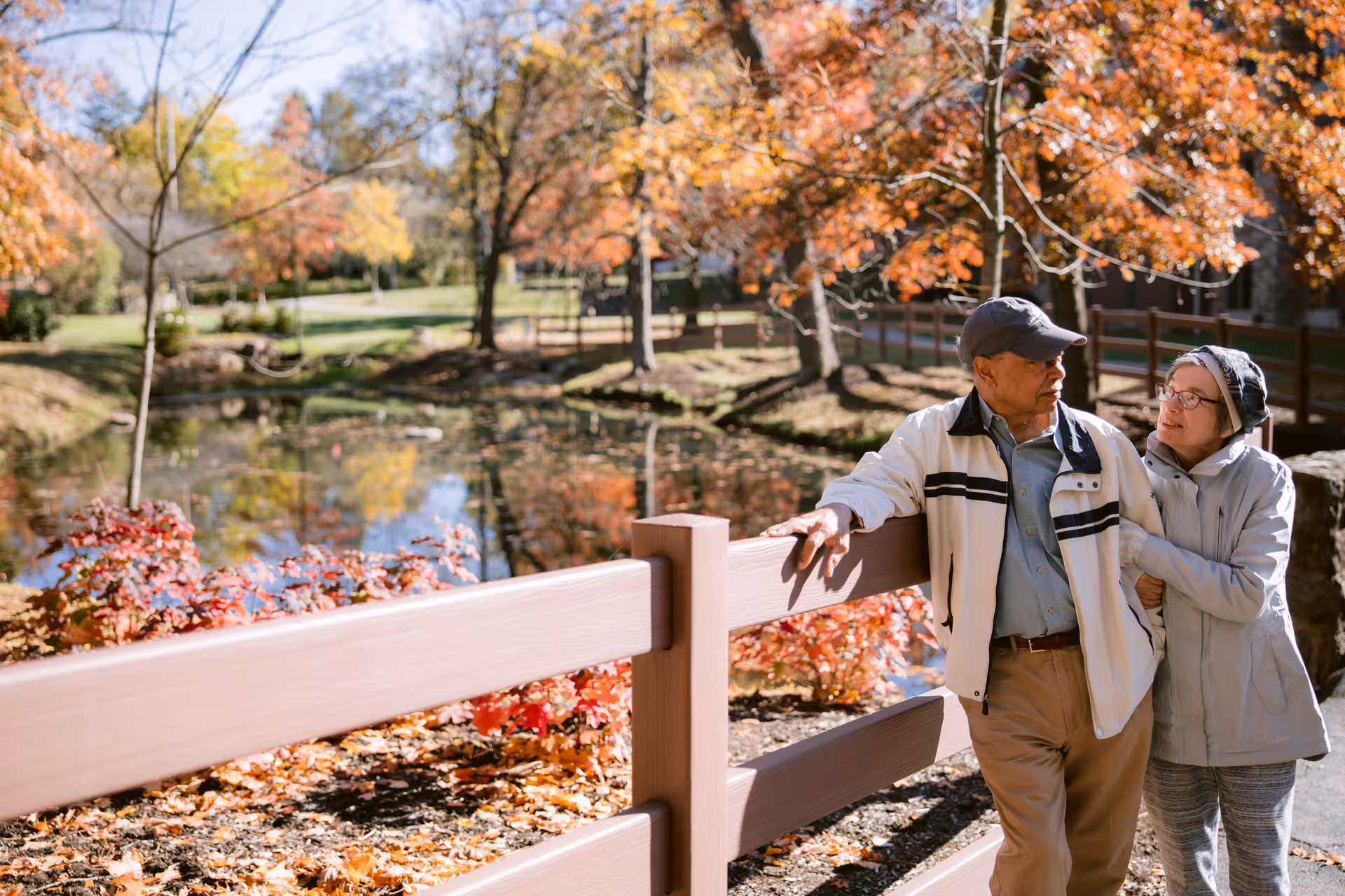 Two older adults walking arm-in-arm along a wooden fence by a pond surrounded by autumn trees.