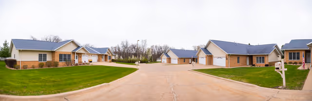 A wide view of a residential senior living community with multiple single-story brick cottages featuring gray roofs, surrounded by green lawns and a paved road in the center under a cloudy sky.