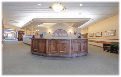 Reception desk area inside The Pavilion Senior Living at Lebanon with wooden paneling, a sign reading 'The Pavilion' on the wall behind the desk, beige walls, carpeted floor, framed pictures on the wall, and a seating area with chairs and a plant.