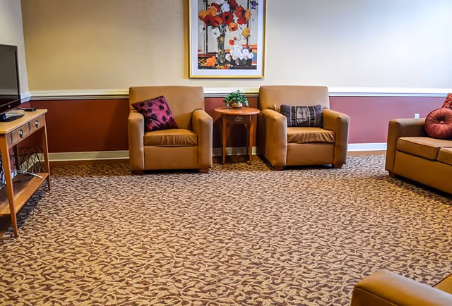 A cozy living room area with two tan armchairs and a tan sofa, each adorned with decorative pillows. A small wooden side table with a plant sits between the armchairs. A framed floral painting hangs on the wall above the chairs. The room has patterned carpet and a two-tone wall with beige and reddish-brown colors.