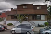 Two-story brown residential building with a covered front porch, exterior stairs and handrails, and cars parked on the street in front.