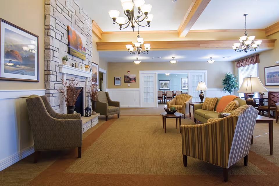 A cozy living room area in a senior living facility featuring a stone fireplace with a mantle decorated with small plants and artwork above it. The room has multiple armchairs and sofas arranged around a coffee table, warm lighting from chandeliers and table lamps, framed paintings on the walls, and a carpeted floor. In the background, there is a dining area visible through open double doors.