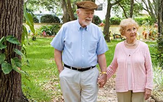An elderly couple holding hands and walking outdoors on a garden path surrounded by trees and greenery. The man is wearing a light blue shirt, beige pants, and a brown cap, while the woman is wearing a pink cardigan and beige pants.