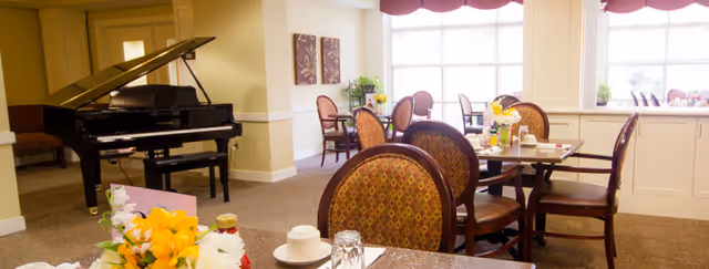 Dining room with round tables and upholstered patterned chairs set with place settings and a grand piano near the windows.