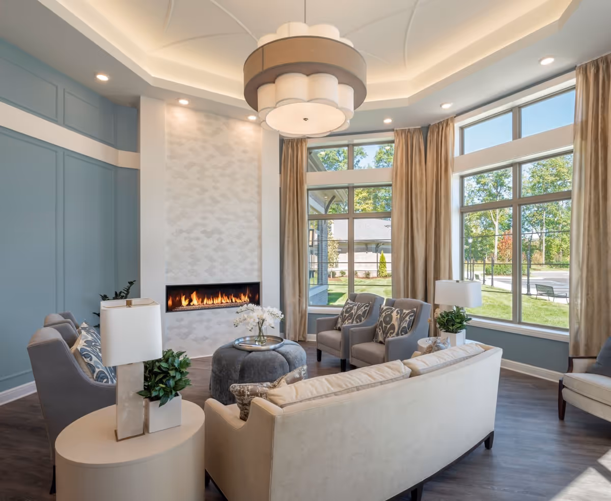 A bright and elegant living room with large windows showing a green outdoor view. The room features a modern fireplace with a white tiled surround, a beige sofa, four gray armchairs with patterned cushions, a round ottoman with a tray and flowers, two side tables with lamps and plants, and a large decorative ceiling light fixture.