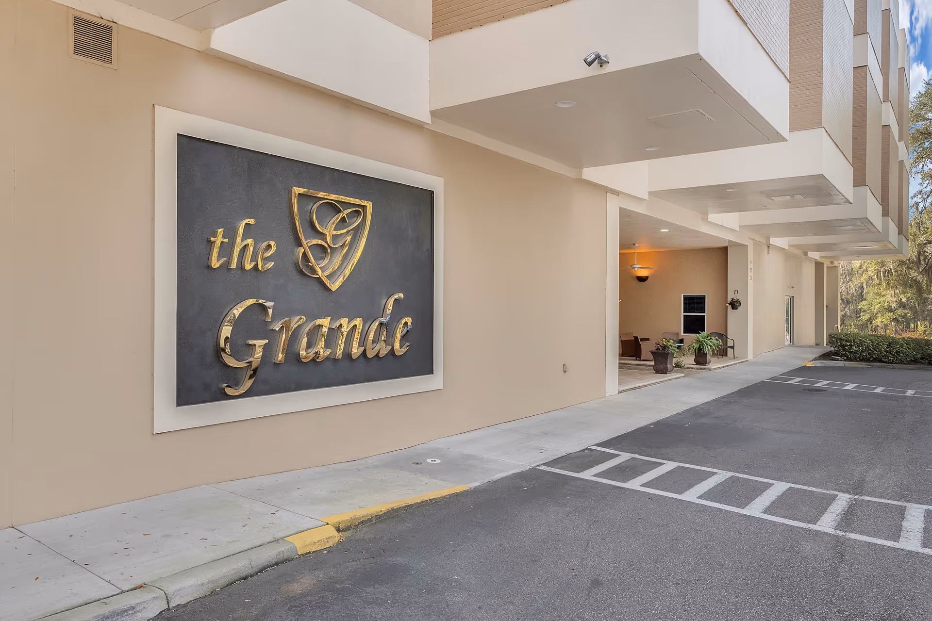Exterior view of a building with a large sign that reads 'the Grande' in gold letters mounted on a beige wall. There is a covered entrance area with some potted plants and chairs, and a parking area with marked spaces in front.