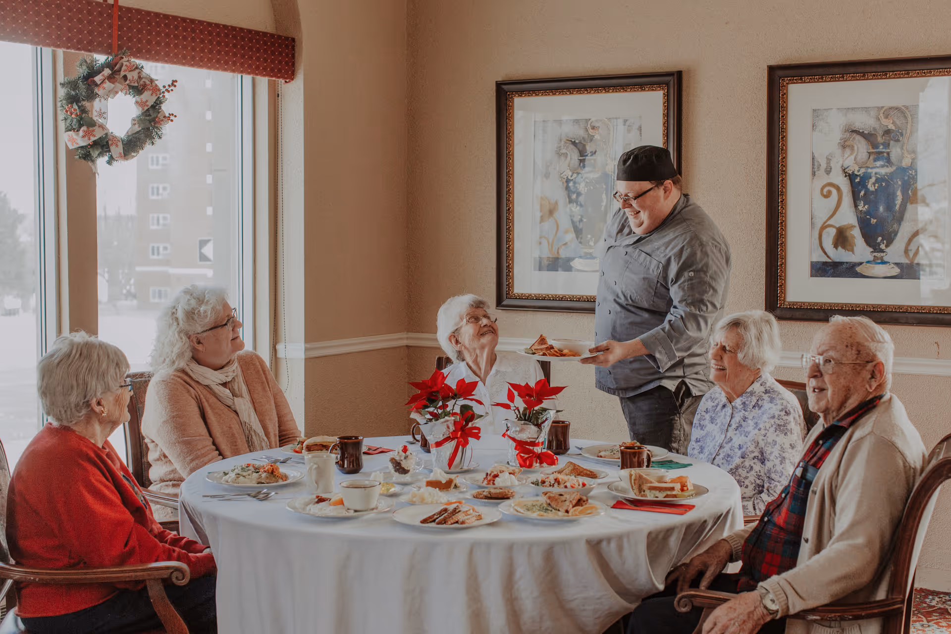 A group of five elderly people sitting around a round dining table with white tablecloth, enjoying a meal together. A chef is standing and serving food to one of the elderly women. The room has beige walls with two framed pictures and a window decorated with a Christmas wreath.