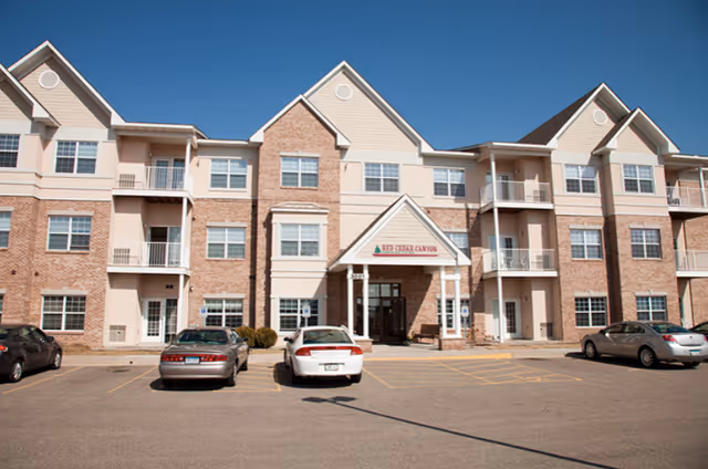 Front facade of a three-story senior living building with a covered entrance, balconies, and parked cars in the driveway.