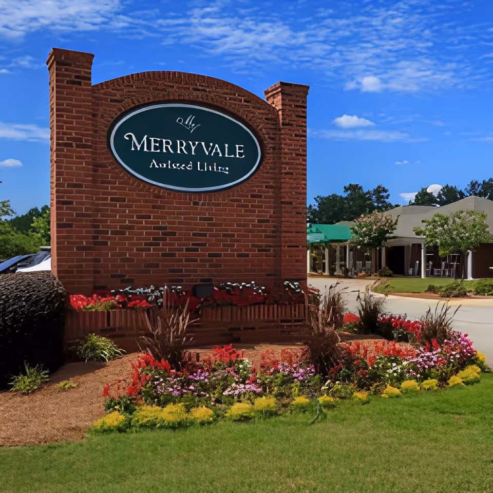 Brick entrance sign reading "MERRYVALE Assisted Living" with landscaped flowerbeds and the facility building visible behind it.