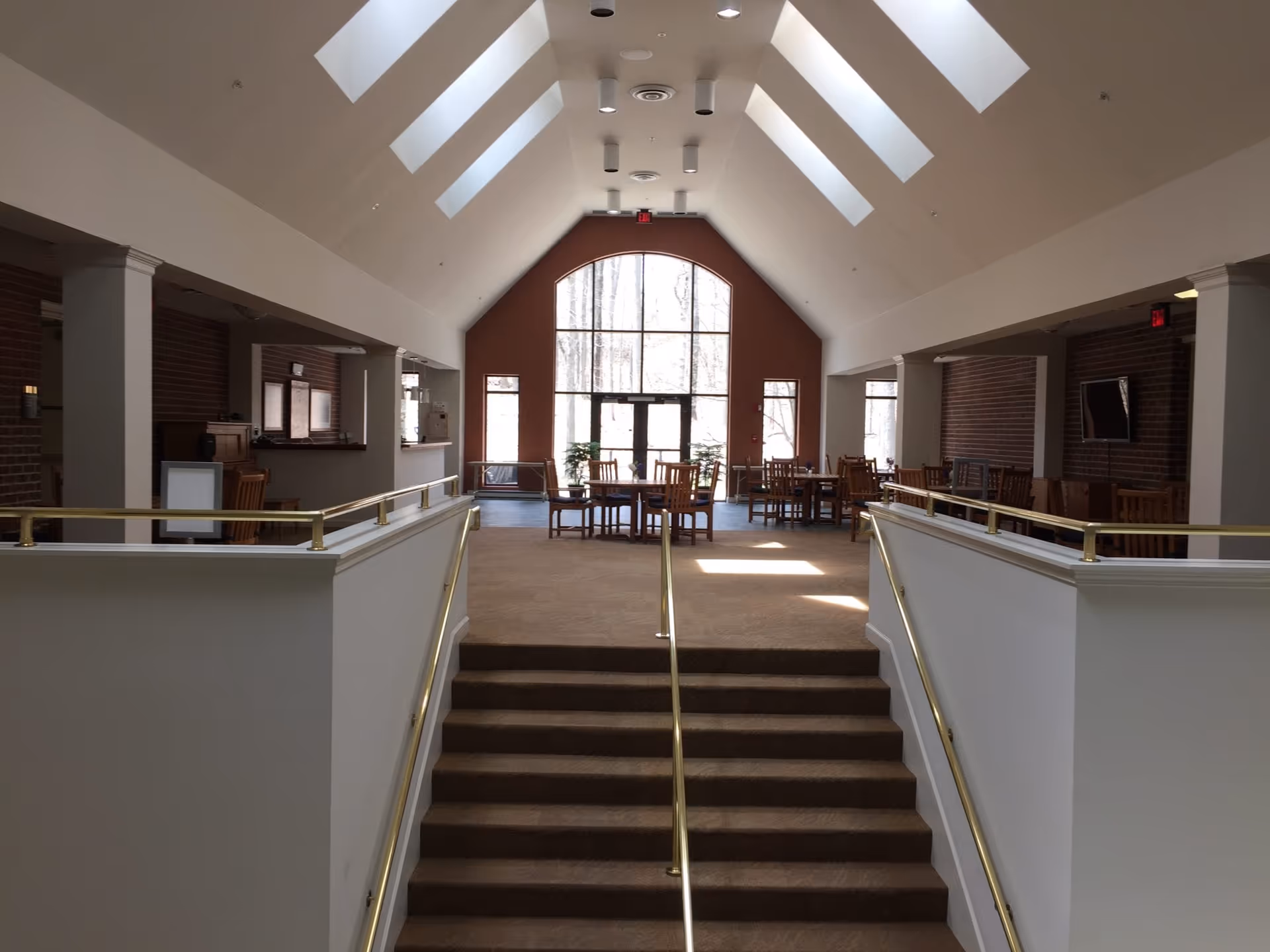 Interior view of a senior living facility common area with a high vaulted ceiling featuring skylights. There are stairs with brass handrails leading up to a carpeted area with wooden tables and chairs. Large windows at the far end let in natural light, and there are brick walls on the sides with a mounted TV on the right.