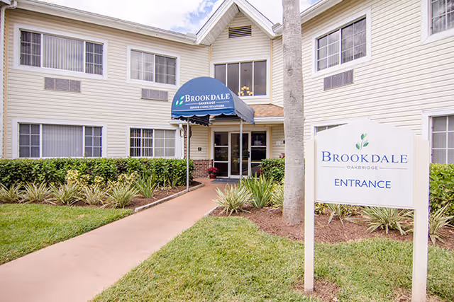 Exterior view of a two-story senior living facility building with beige siding and multiple windows. A blue awning above the entrance reads 'Brookdale Oakbridge Senior Living Solutions.' A sidewalk leads to the entrance, flanked by green grass and landscaped plants. A white sign near the sidewalk says 'Brookdale Oakbridge Entrance.'