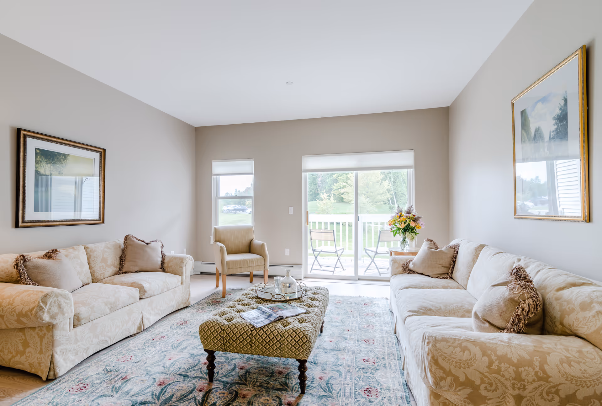 A bright living room with two beige patterned sofas facing each other, a cushioned ottoman in the center with a tray and magazines on it, a single armchair near a window, and a sliding glass door leading to an outdoor patio with two chairs and a small table. The walls are light-colored with framed artwork, and a floral arrangement is on a side table next to one sofa.