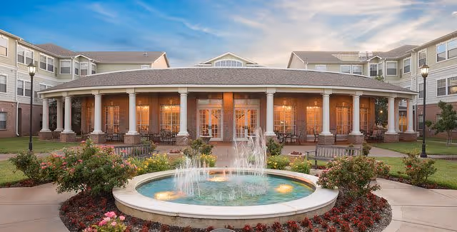 Exterior view of a senior living facility with a central circular water fountain surrounded by flowers and benches. The building has a covered porch with columns and warm lighting inside, under a partly cloudy sky at dusk.