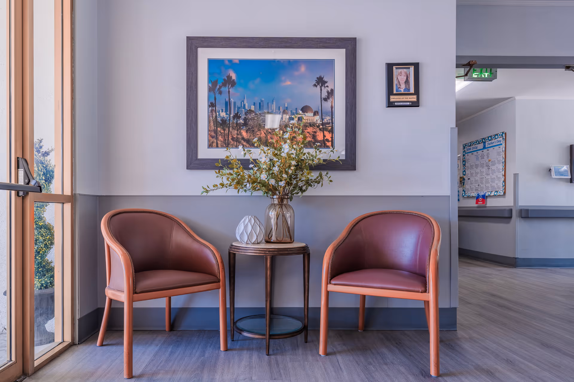 Two leather chairs flank a small round table with a vase of flowers beneath framed artwork in a healthcare facility waiting area.