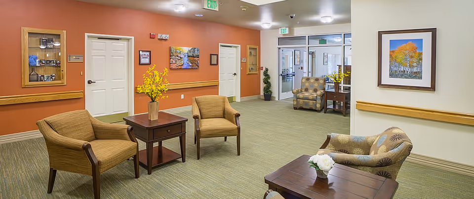 A well-lit senior living facility common area with comfortable armchairs arranged around wooden tables. The walls are decorated with framed artwork and a display cabinet. There are exit signs above doors leading to other rooms and hallways.