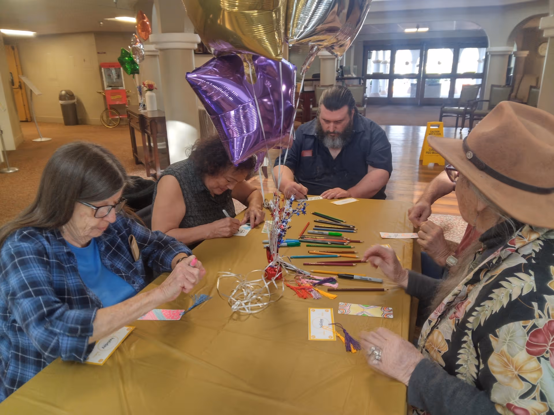 A group of elderly people sitting around a table covered with a yellow tablecloth, engaged in a craft activity with colored pencils and markers. There are star-shaped balloons in purple, gold, and silver tied to a centerpiece on the table. The setting appears to be a common area inside a senior living facility with large windows and an open entrance in the background.
