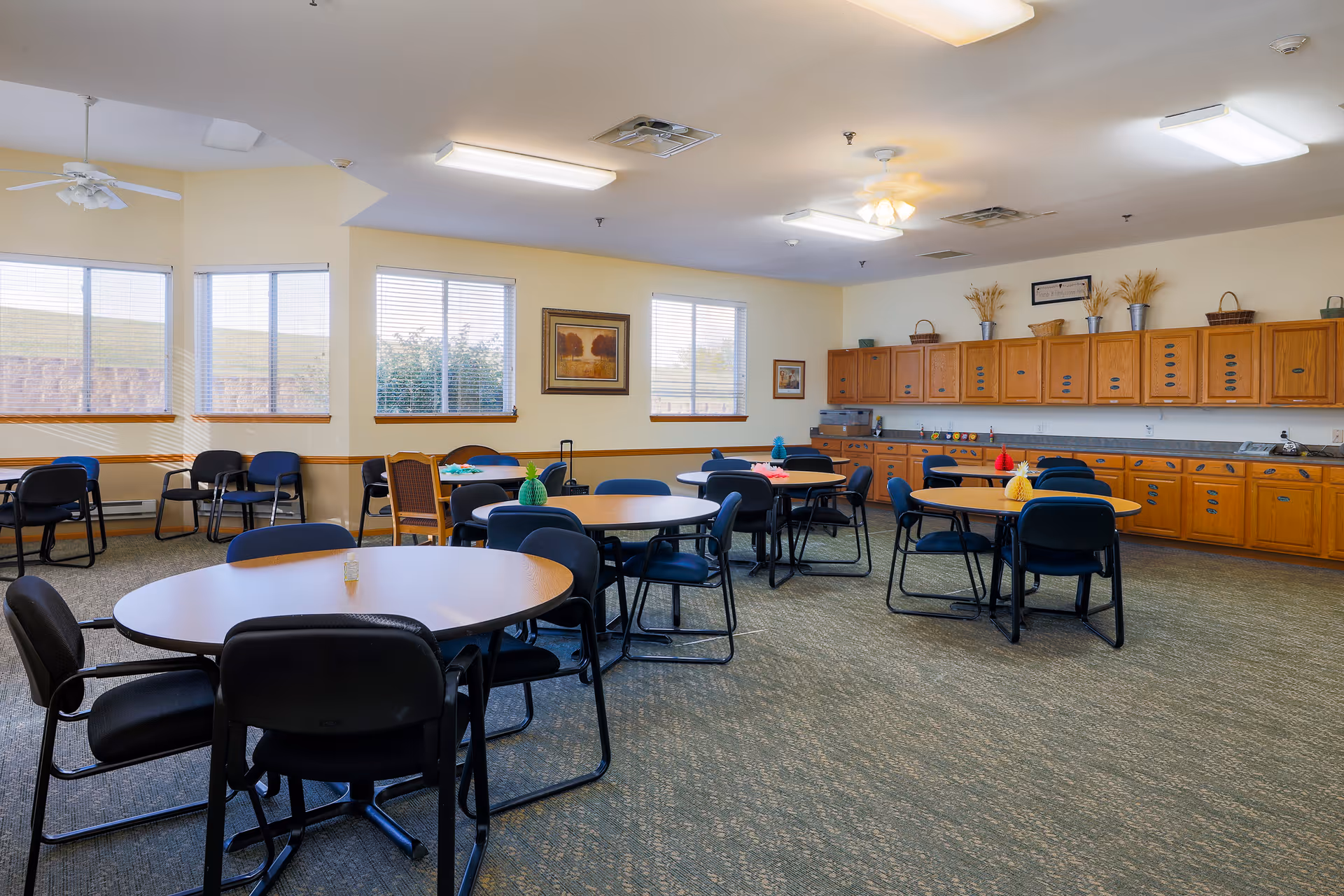 Bright dining/activity room with several round tables and chairs, wood cabinets along one wall and windows letting in natural light.