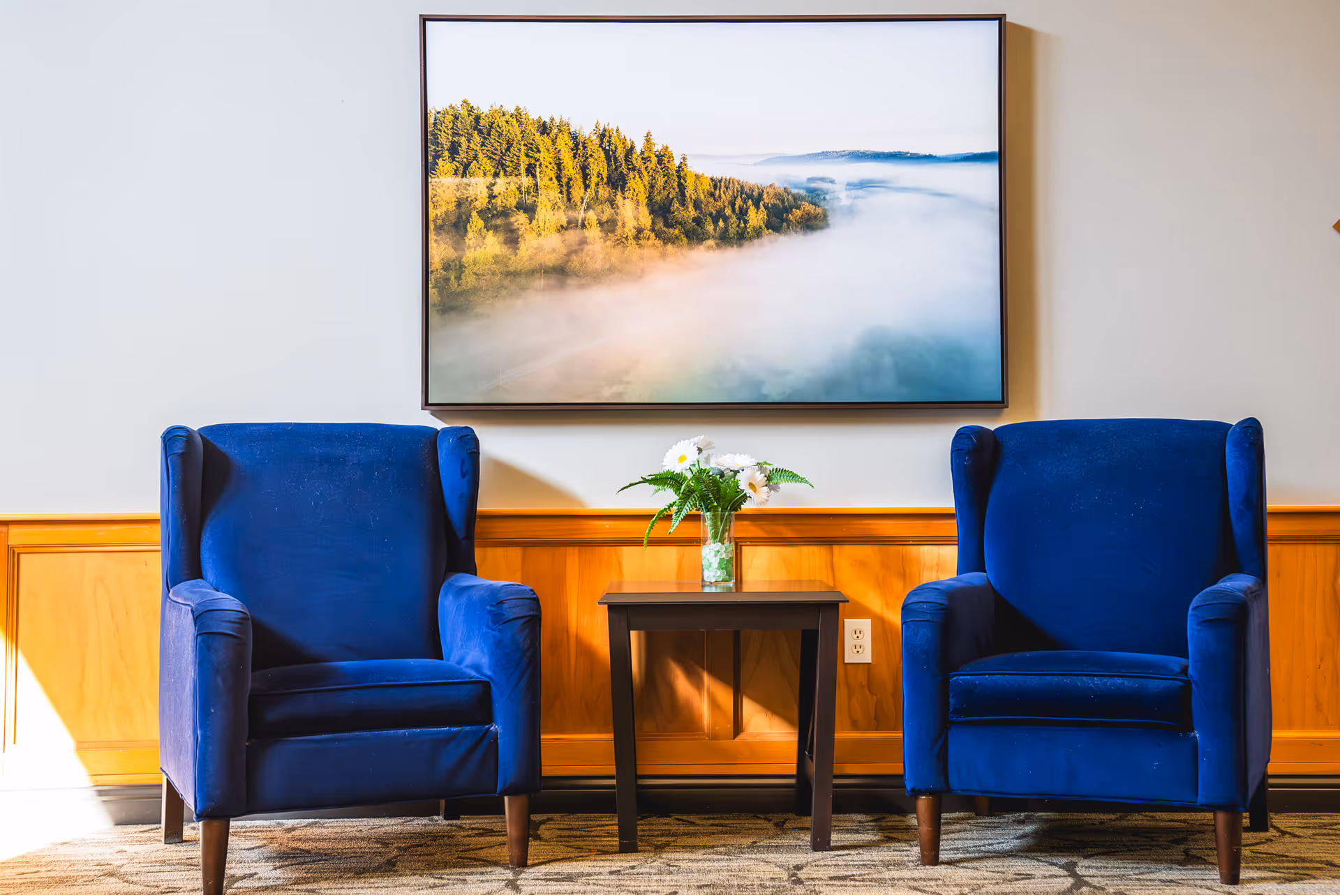 Two blue upholstered armchairs with wooden legs are positioned on either side of a small dark wooden table holding a vase with white flowers and green leaves. Behind the chairs is a wooden wainscoting on the wall, above which hangs a large framed photograph of a misty forest landscape.