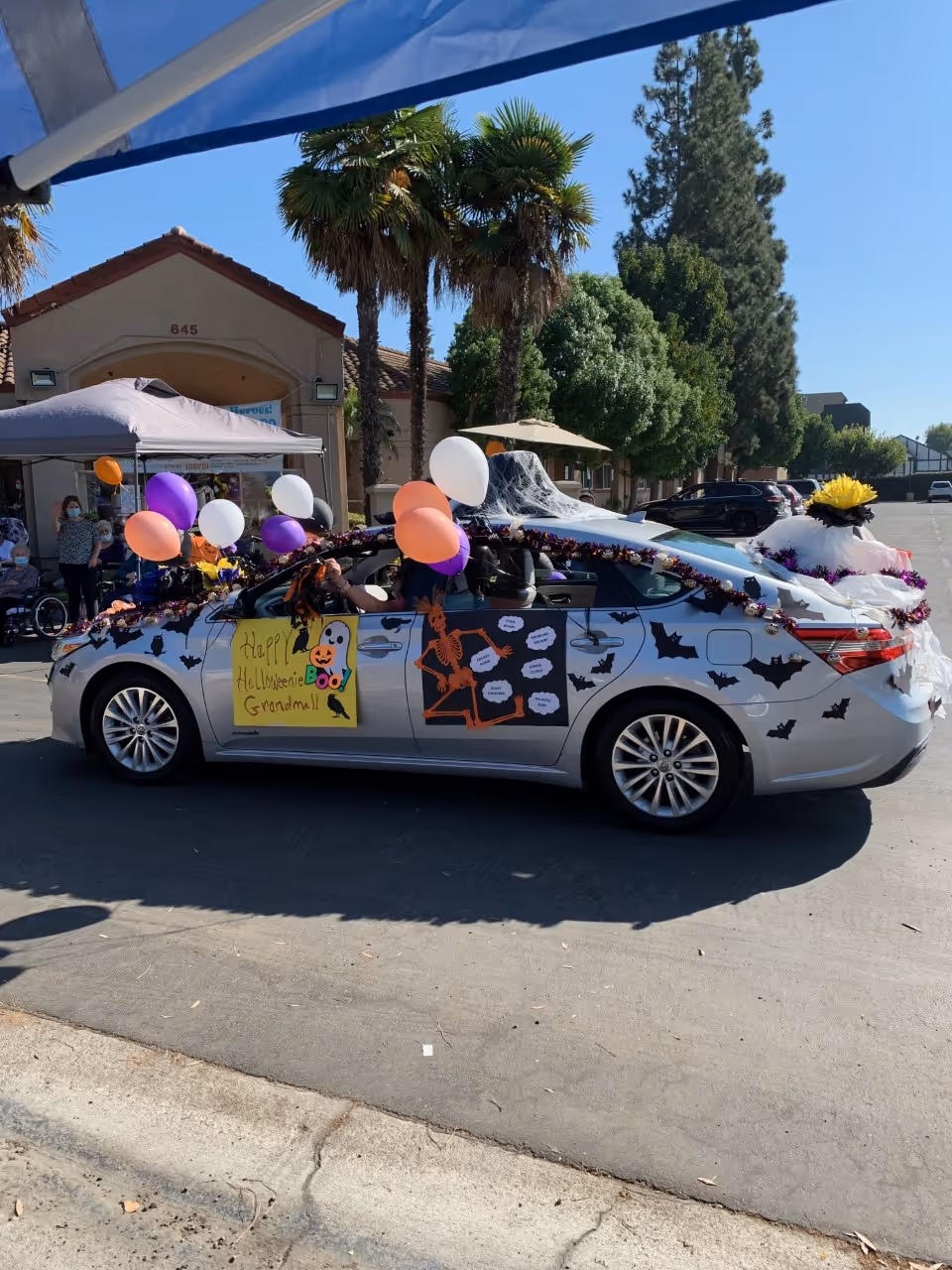 A silver car decorated with Halloween-themed decorations including balloons, paper bats, a skeleton cutout, and a sign that says 'Happy Halloween Grandma!!'. The car is parked outside a building with palm trees and people standing under a canopy in the background.