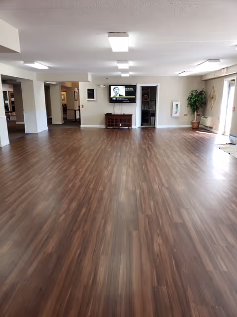 Spacious empty multipurpose room with wood-look flooring, a wall-mounted TV and console at the far end, and windows with a potted plant to the right.