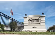 Outdoor view of a stone sign for Avante at Melbourne Rehabilitation Center with the address 1420. The sign is situated near a black metal fence and an American flag is visible in the background under a blue sky.