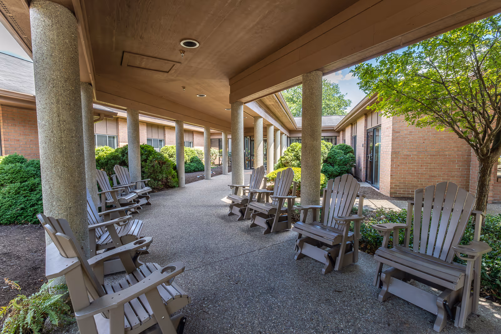 Covered outdoor walkway at Elison Assisted Living & Memory Care of Marietta with multiple wooden Adirondack chairs arranged along the sides. The walkway is supported by large concrete columns and surrounded by green bushes and trees, with brick building walls visible on both sides.