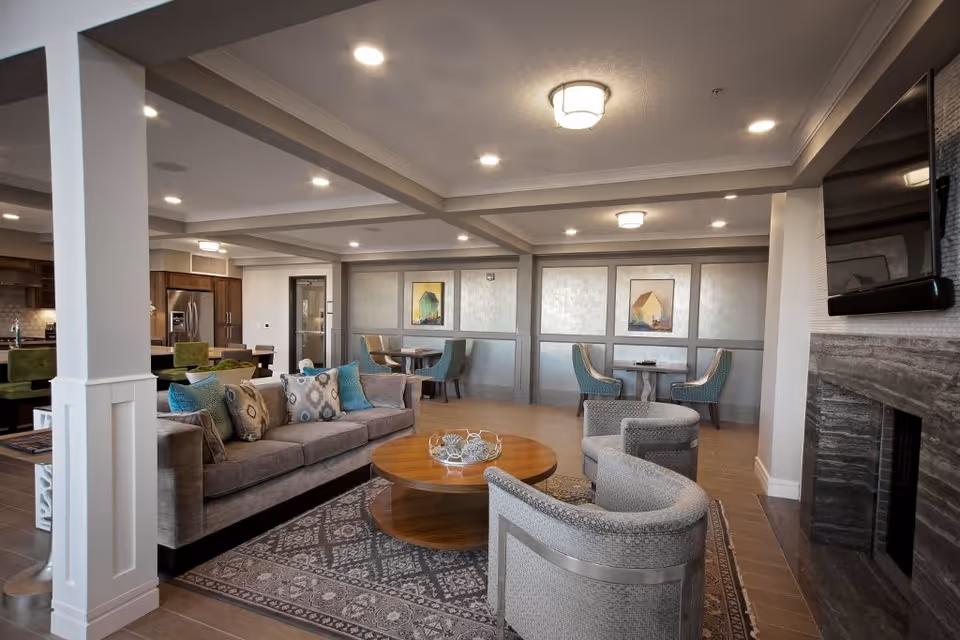 A spacious and well-lit living room area in a senior living facility featuring a gray sofa with patterned cushions, two gray armchairs, a round wooden coffee table on a patterned rug, and a fireplace with a mounted flat-screen TV. In the background, there are dining tables with teal chairs and a kitchen area with wooden cabinets and stainless steel appliances.