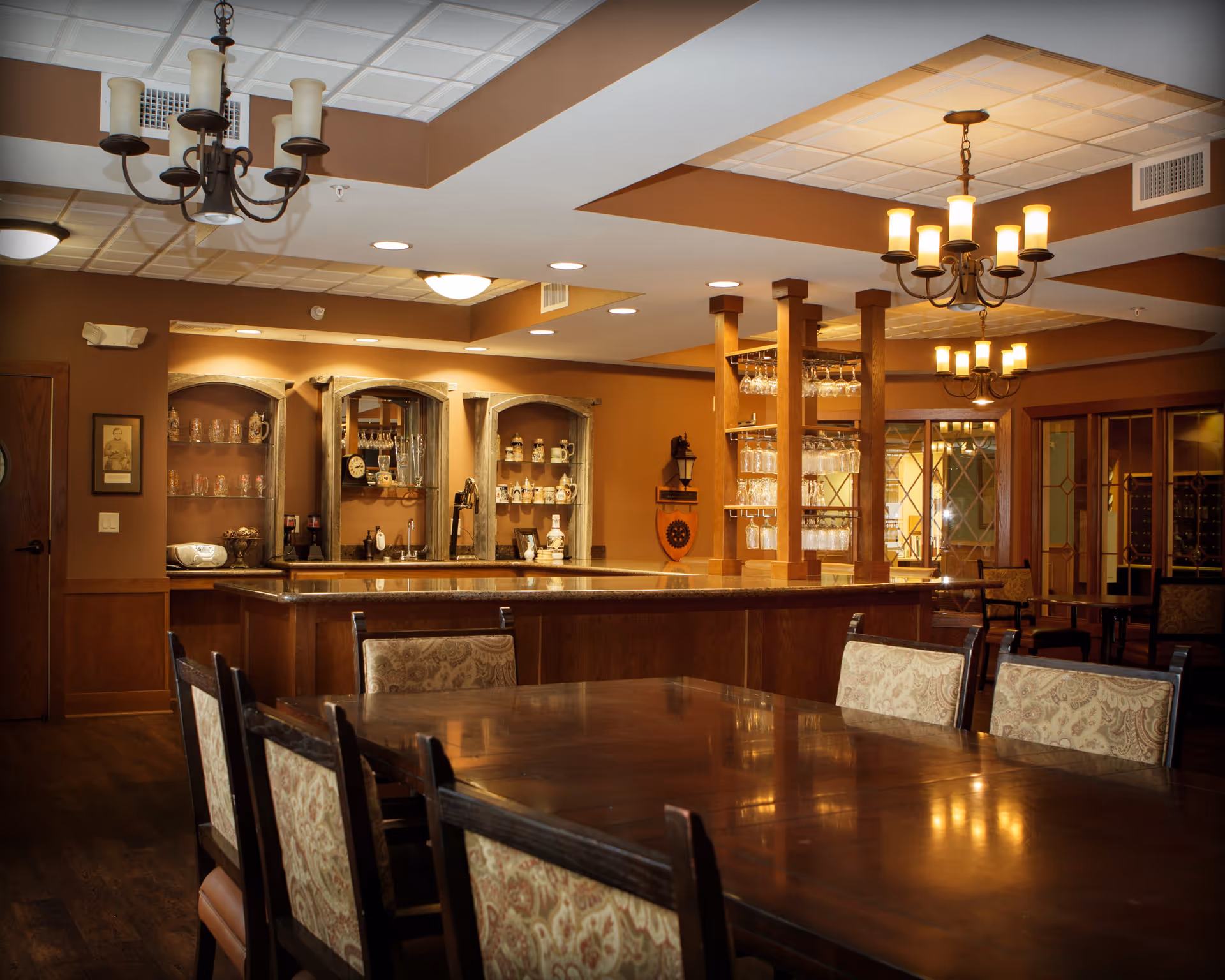 A warm, inviting dining area with a large wooden table surrounded by upholstered chairs. In the background, there is a bar with shelves holding glassware and decorative items. The room features warm lighting with chandeliers hanging from the ceiling and wood-paneled walls.