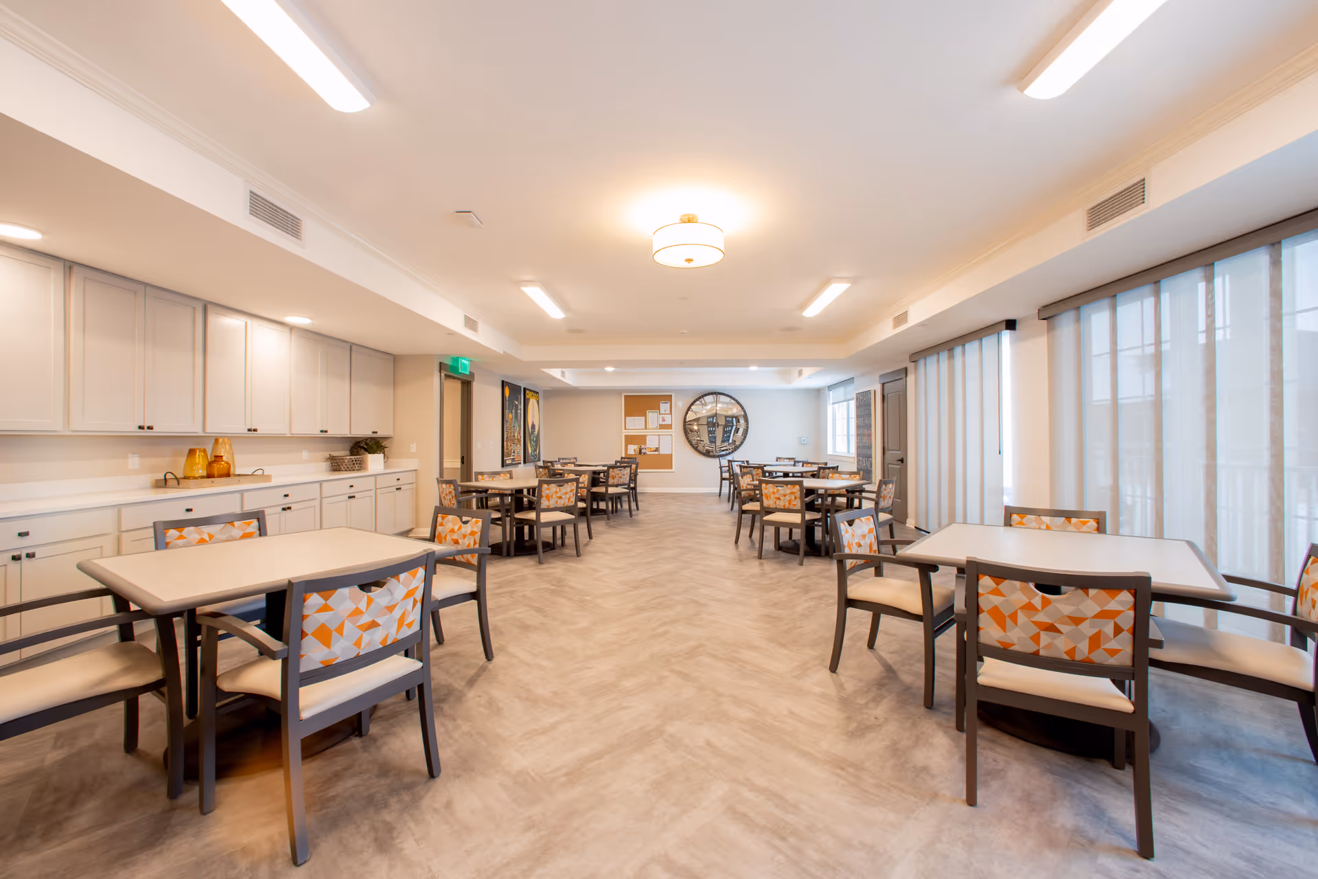 A spacious and well-lit dining room in a senior living facility with multiple tables and chairs arranged neatly. The chairs have patterned cushions with orange and beige geometric designs. There are large windows with sheer curtains on the right side, and cabinets with countertop space on the left. The room has a light-colored floor and ceiling lights providing ample illumination.