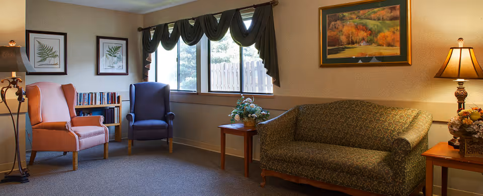 A cozy sitting area in a nursing facility with two armchairs, a patterned sofa, two side tables with lamps and floral arrangements, framed botanical prints on the wall, and a window with dark green curtains letting in natural light.