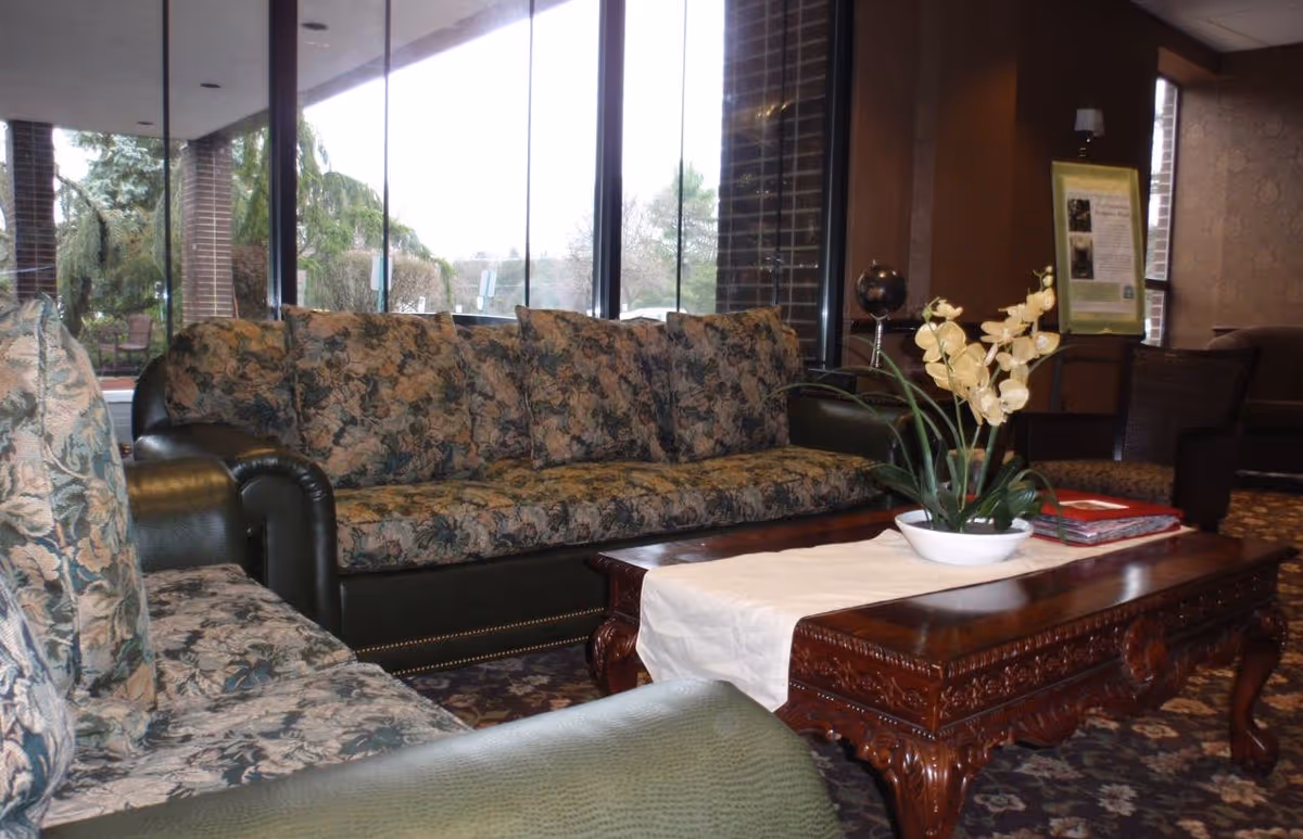 A cozy sitting area with floral patterned sofas and a carved wooden coffee table with a white table runner and a potted plant. Large windows in the background show trees and outdoor scenery, and the room has warm brown walls and carpeted flooring.