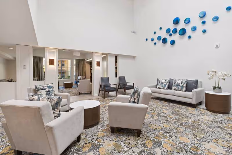A bright and spacious living room area with high ceilings, featuring a beige sofa with patterned cushions, four beige armchairs with matching cushions, two dark wooden chairs, a round coffee table, and a side table with a white orchid plant. The room has a patterned carpet and a wall decorated with blue circular art pieces.
