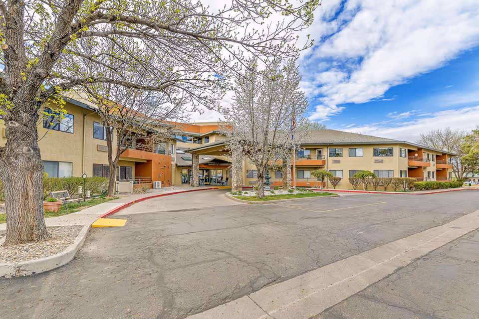 Exterior view of Solstice Senior Living at Mesa View, showing a two-story building with beige and orange walls, surrounded by trees with budding leaves and a partly cloudy sky. The entrance has a covered driveway with a circular drop-off area and some landscaping.