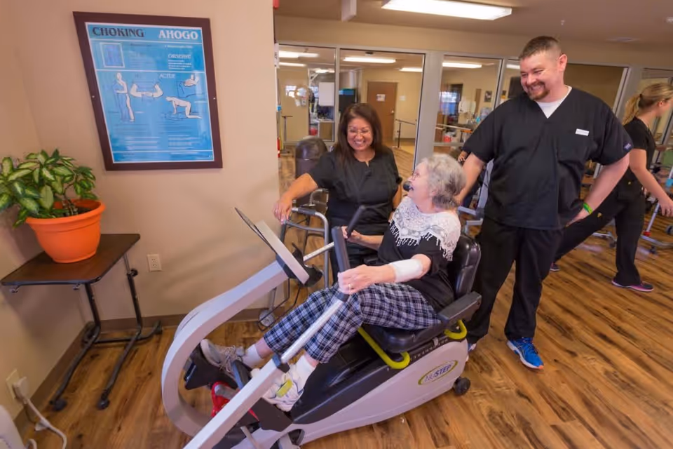 An elderly woman using a NuStep exercise machine in a rehabilitation facility, assisted by two healthcare staff members dressed in black scrubs. The room has wooden flooring, a plant on a small table, and a framed poster about choking on the wall.