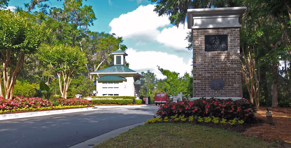 Entrance to The Marshes of Skidaway Island featuring a brick sign with the facility name and number 95, surrounded by landscaped flowers and trees, with a guardhouse and a red vehicle in the background under a partly cloudy sky.