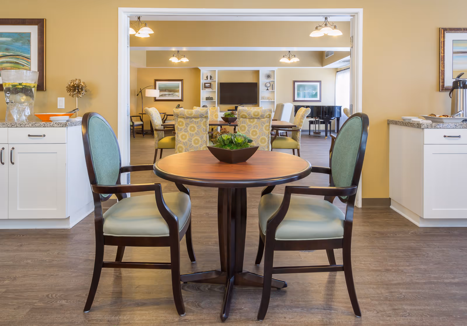 A cozy seating area in an assisted living facility featuring a round wooden table with two green cushioned chairs. In the background, there is a larger dining table surrounded by floral-patterned chairs, a flat-screen TV mounted on a white shelving unit, framed artwork on the walls, and a black grand piano near a window with sheer curtains. The room has warm yellow walls and wood flooring, with countertops on either side holding a water dispenser and coffee setup.