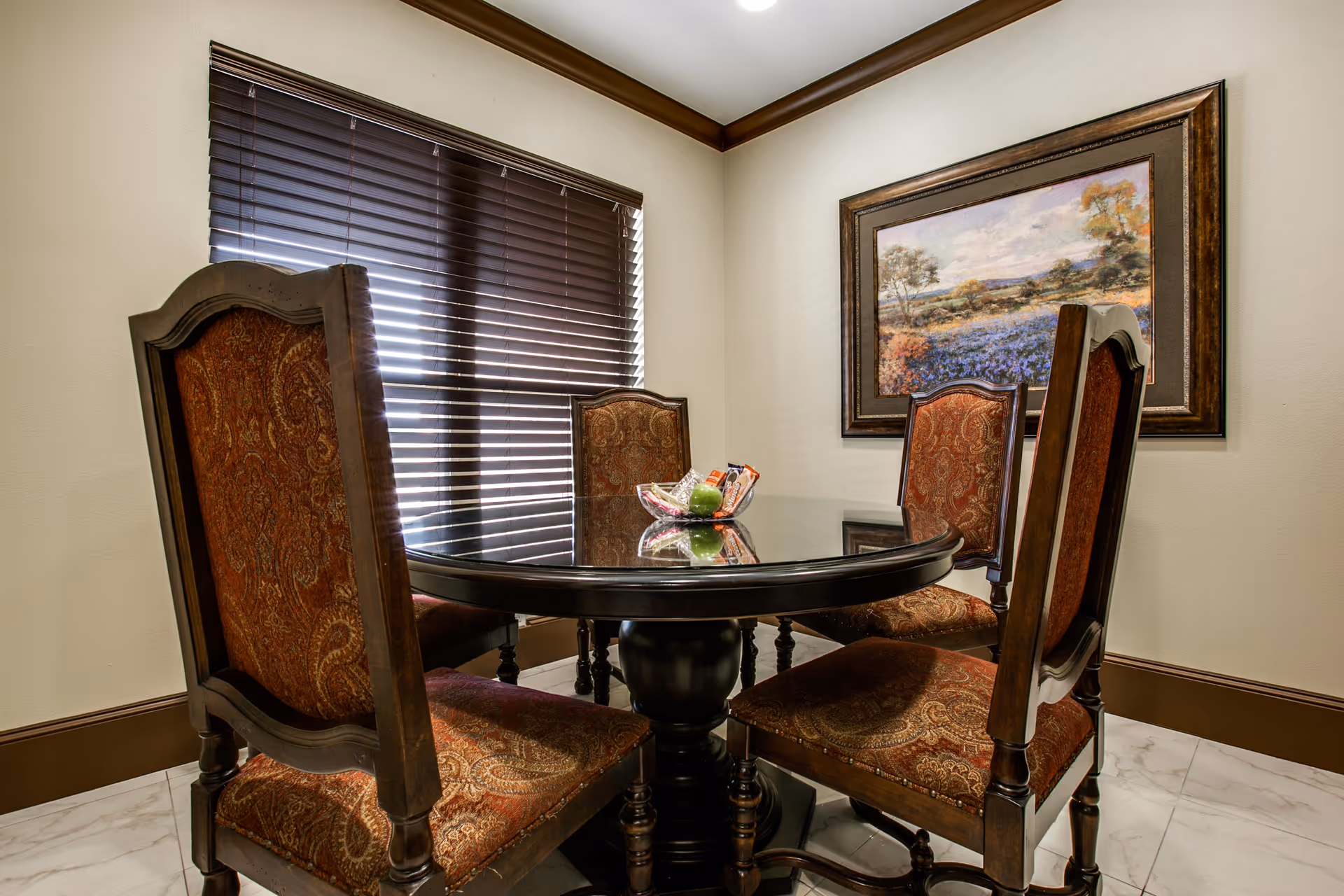 A small dining area with a round dark wooden table surrounded by four ornate wooden chairs with patterned upholstery. A window with closed blinds is on one wall, and a framed landscape painting hangs on another wall. The floor is tiled with a light marble pattern.
