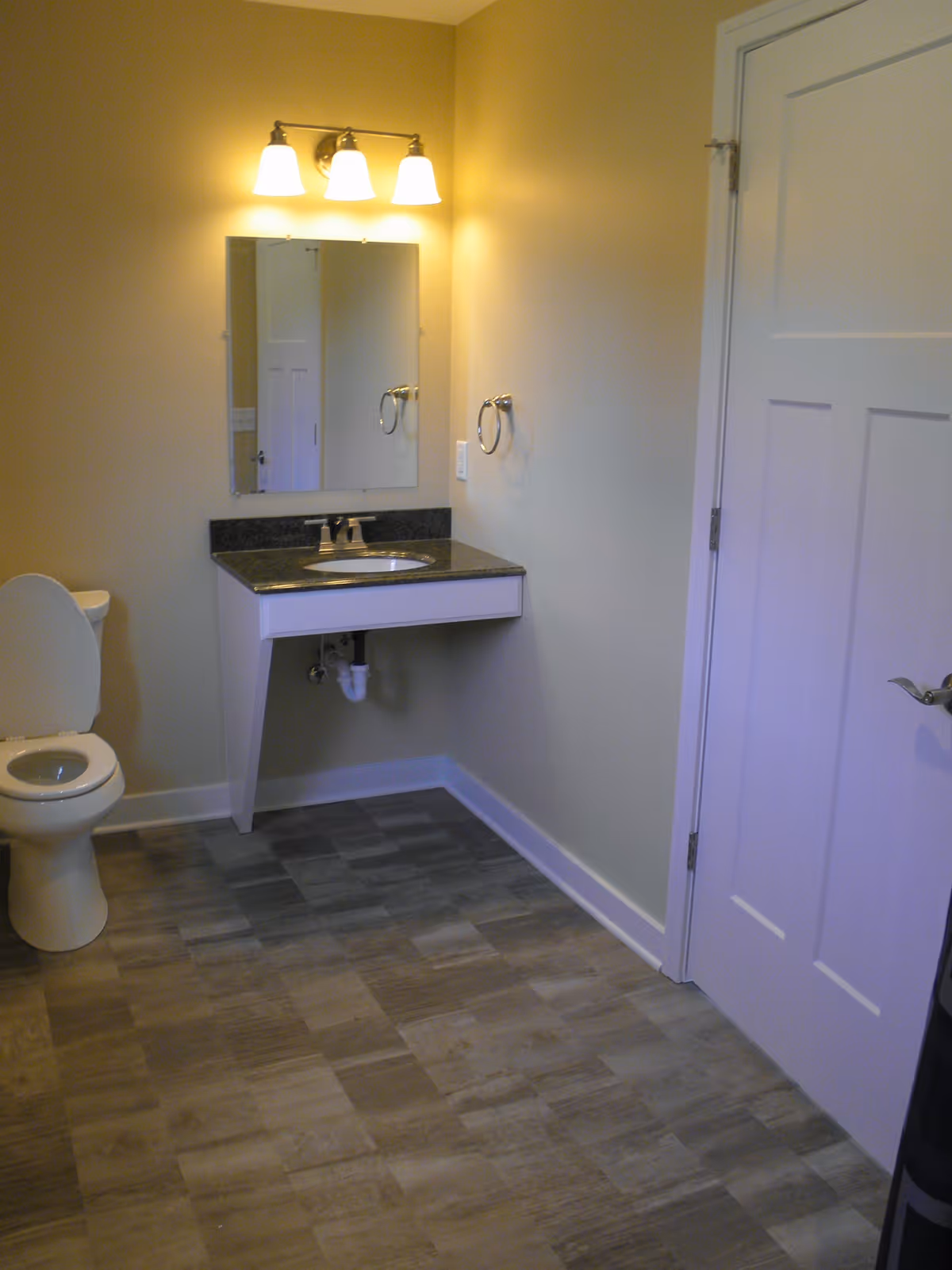 A clean bathroom with a white toilet, a wall-mounted sink with a black countertop, a mirror above the sink, and a three-light fixture mounted on the wall above the mirror. The bathroom has beige walls, a white door, and a tiled floor with a wood-like pattern.
