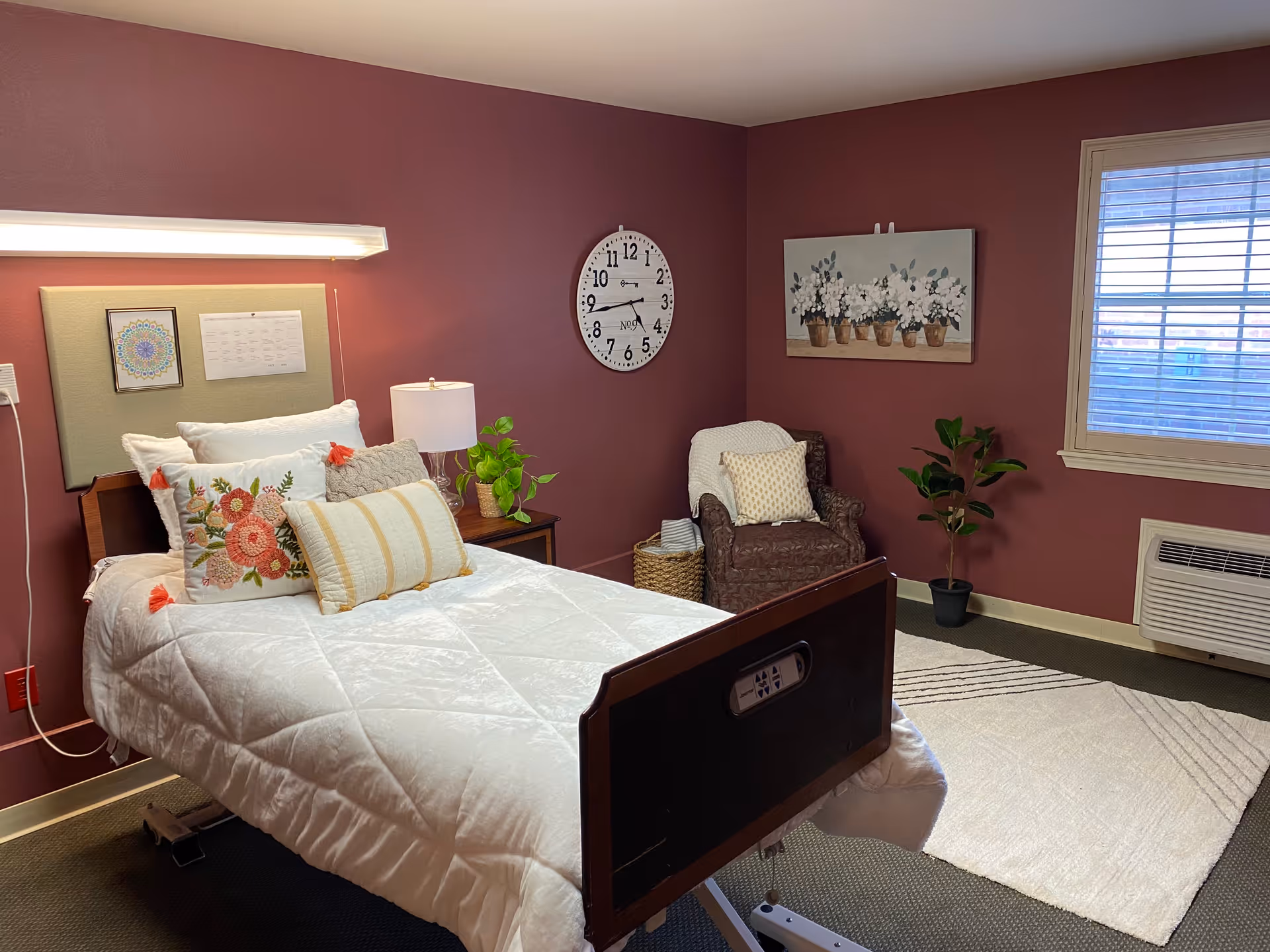 A neatly furnished nursing facility bedroom with a made bed, bedside table and lamp, armchair, wall clock, and window.
